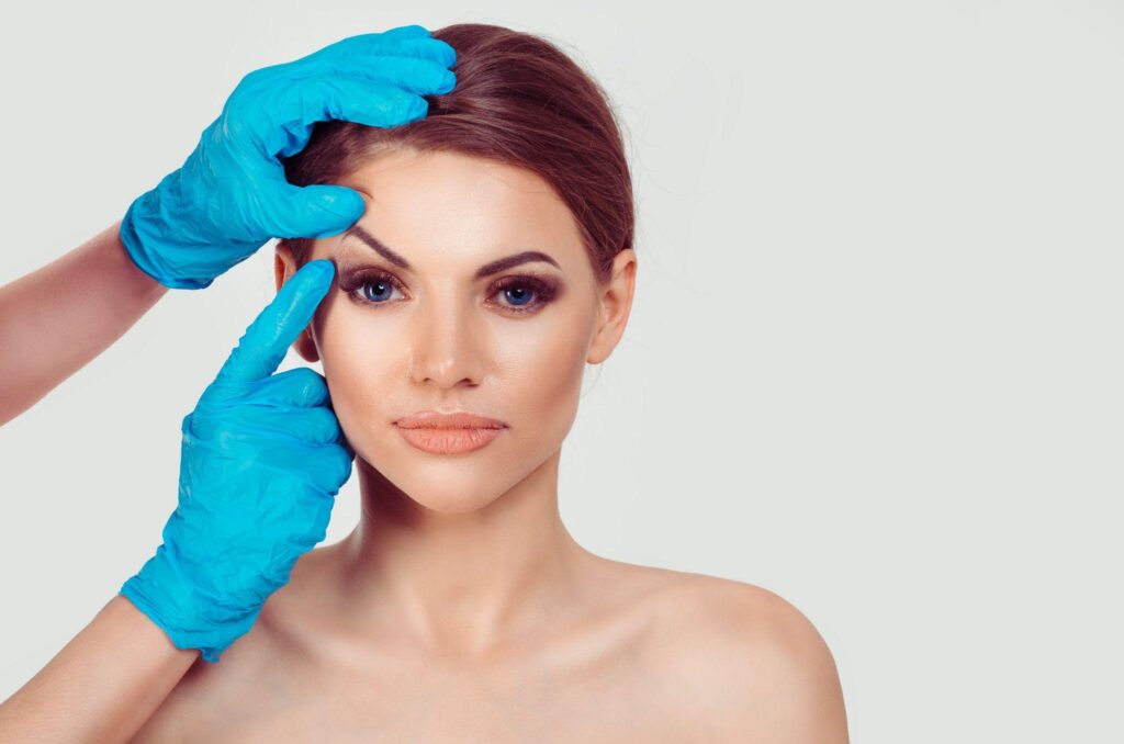 Beautiful middle age woman getting ready for eyelid lift plastic surgery doctor hands in blue gloves point fingers to her eye on white